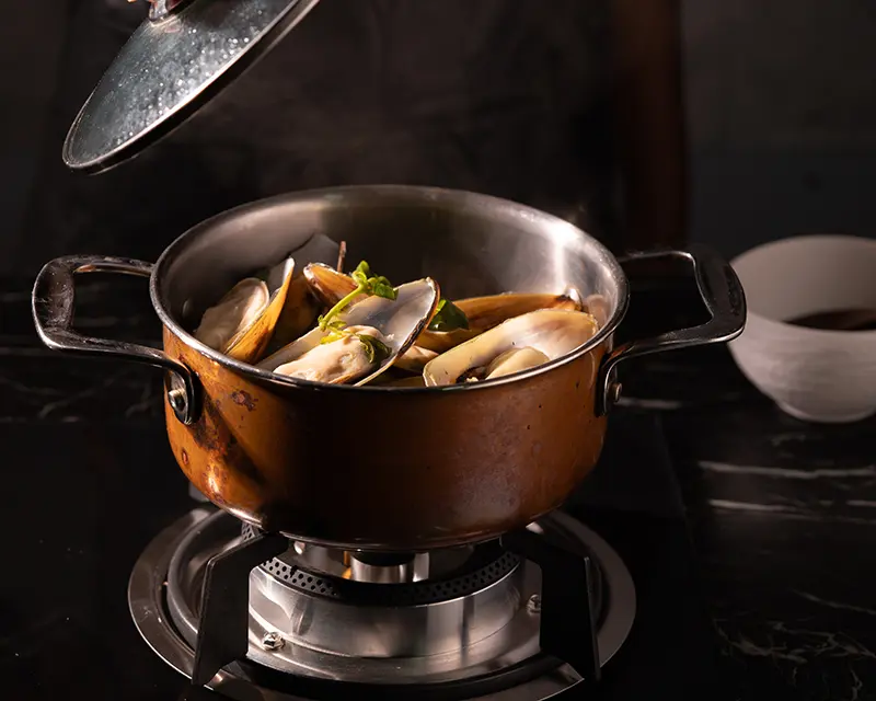 Mussels steaming in a copper pot on the stove, with their shells open as the lid is lifted to reveal the freshly cooked mussels.