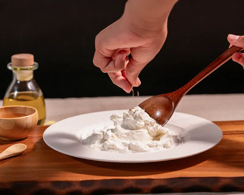 Hand adding salt to yogurt with a spoon, showing how to salt yogurt at the final surface seasoning stage.