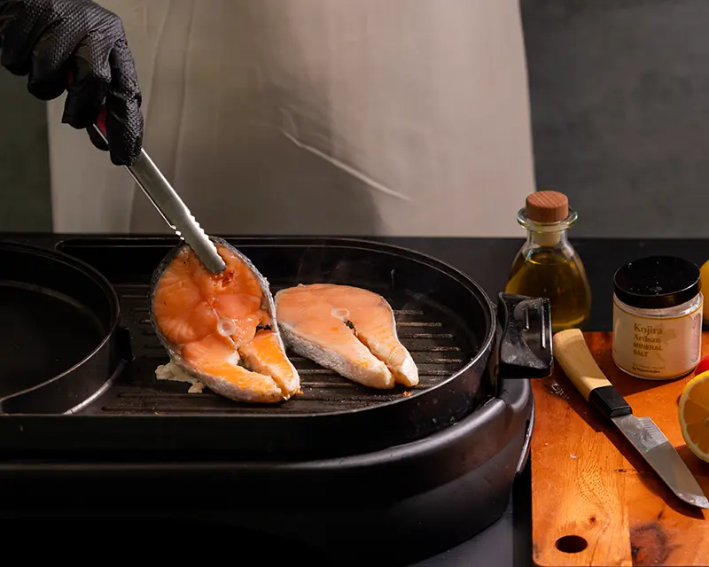 How to season salmon before grilling, shown as a salted salmon steak is placed onto a hot grill beside a second steak.
