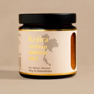 Kojira Artisan Mineral Salt in an amber glass jar with a cream label, gold lettering and a map of Thailand, photographed against a warm beige background.
