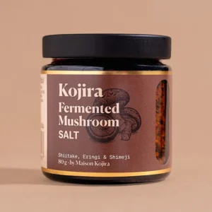 Kojira Fermented Mushroom Salt in an amber glass jar, made with shiitake, eringi and shimeji mushrooms, against a warm beige background.