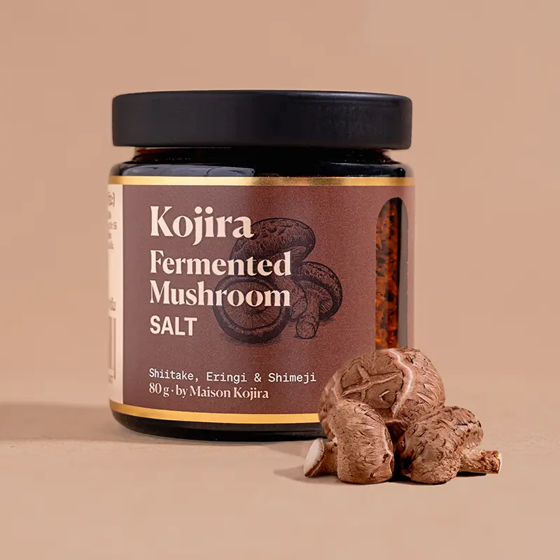Kojira Fermented Mushroom Salt in an 80 g amber glass jar with shiitake mushrooms arranged in front, against a warm beige background.