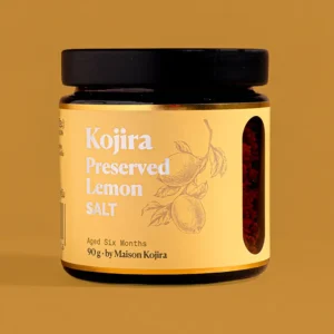 Kojira Preserved Lemon Salt in a 90 g amber glass jar with a golden-yellow label, made with lemons preserved for six months, against an ochre background.