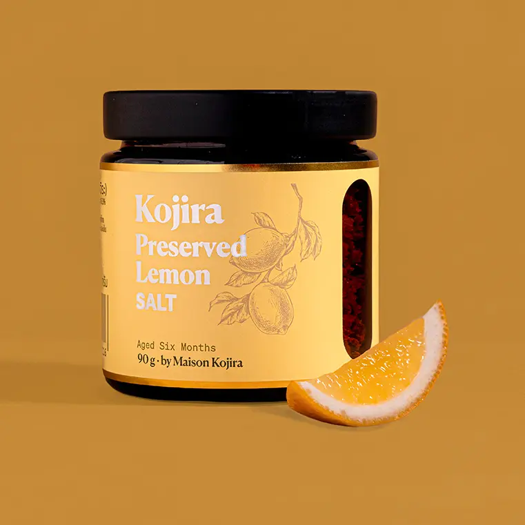 Kojira Preserved Lemon Salt in an amber-glass jar beside a fresh lemon wedge against a warm terracotta background.