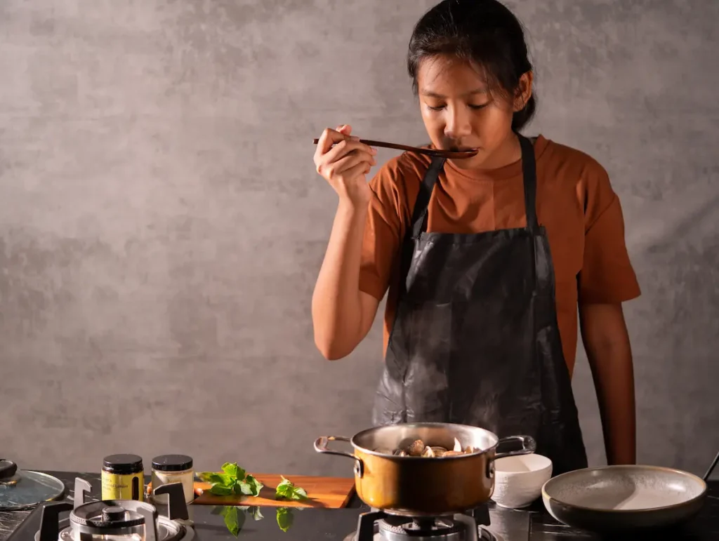 Maison Kojira food press release photograph showing a cook tasting a dish from a wooden spoon, with gourmet salt jars beside the stove.