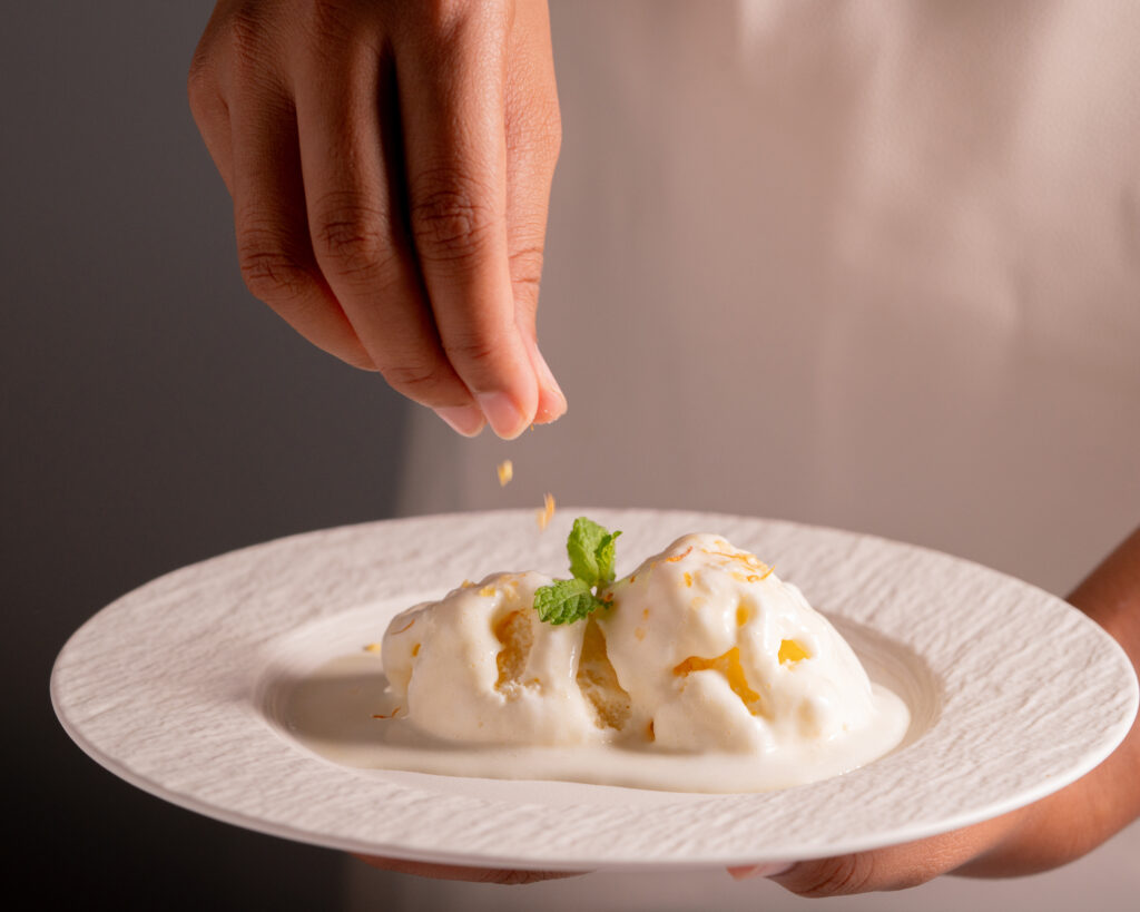 Hand sprinkling finishing salt over ice cream garnished with citrus zest and fresh mint, photographed for Maison Kojira's food press collection.