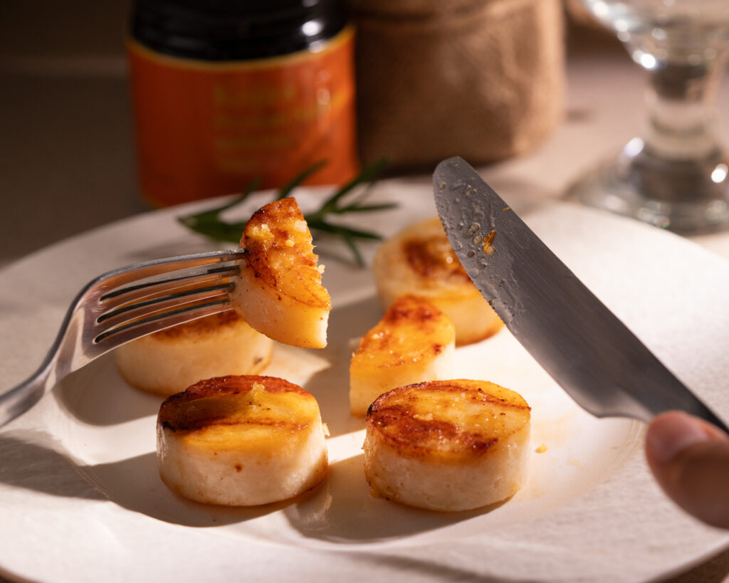 Maison Kojira food press release photograph of golden-seared scallops, with a fork lifting a bite from a white plate.