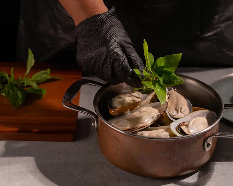 A cook adding fresh basil to half-shell mussels in a copper pot for a mussels recipe with garlic, preserved lemon and no wine.