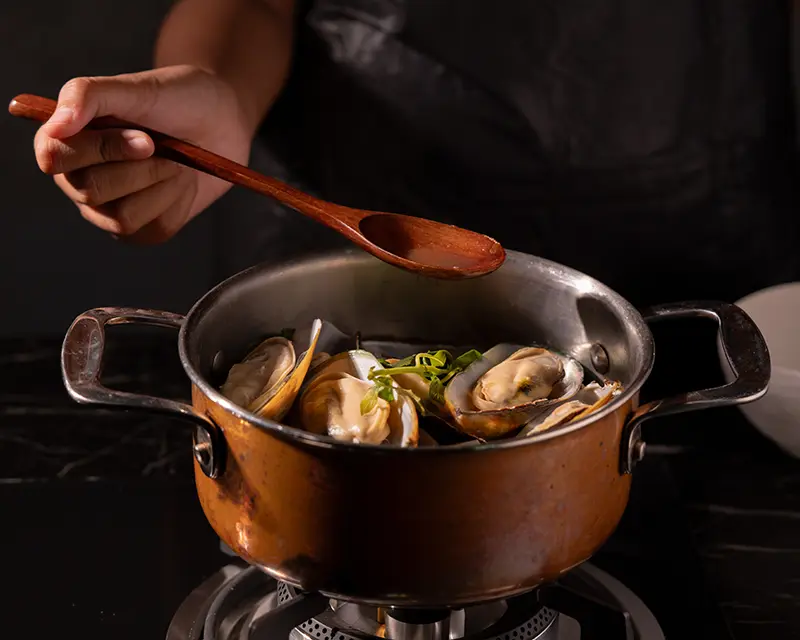 Half-shell mussels cooking in a copper pot on the stove, with a wooden spoon held above the mussels during preparation of a lemon and garlic mussels recipe.