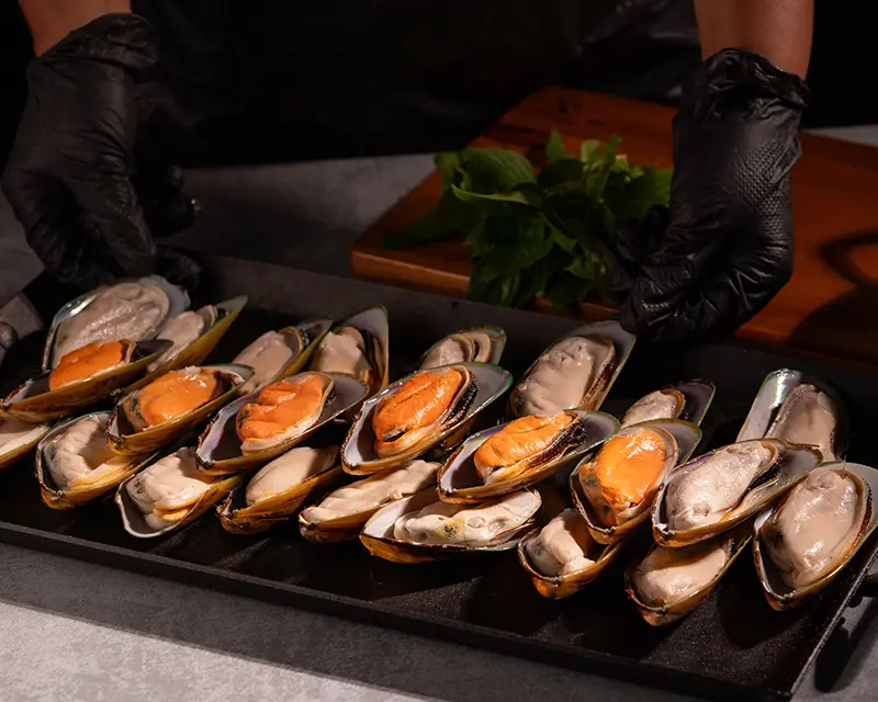 Half-shell mussels arranged on a cooking tray, with gloved hands preparing the shellfish for a lemon and garlic mussels recipe.