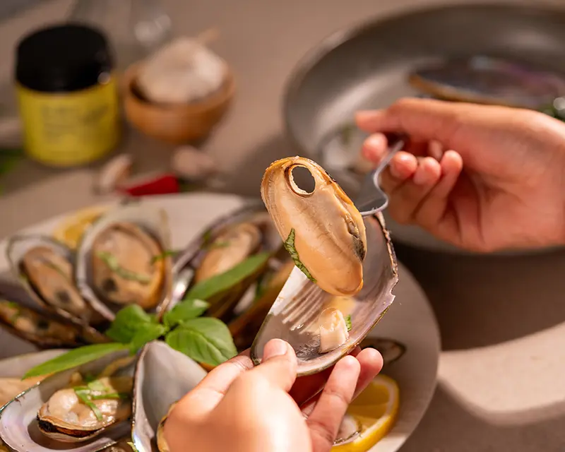 A fork lifting a tender cooked mussel from its half shell over a plate of mussels with fresh basil and lemon slices.