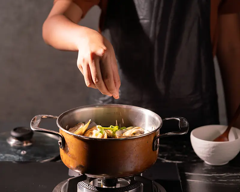 A cook sprinkling seasoning over mussels in a copper pot for a mussels recipe with preserved lemon, garlic and fresh basil.