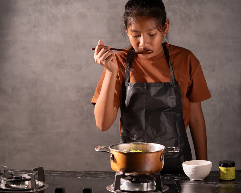 A cook tasting the broth from a wooden spoon while preparing a mussels recipe with preserved lemon, garlic and fresh basil.