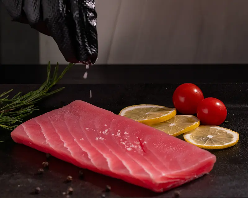 Tuna steak seasoning with mineral salt sprinkled by hand over a raw tuna steak before searing, with lemon and rosemary.
