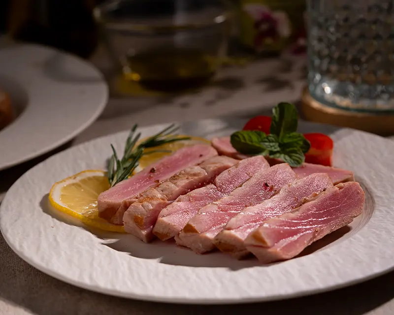 Tuna steak seasoning with salt and pepper on sliced, seared tuna with a pink centre, served with lemon, rosemary and cherry tomatoes.
