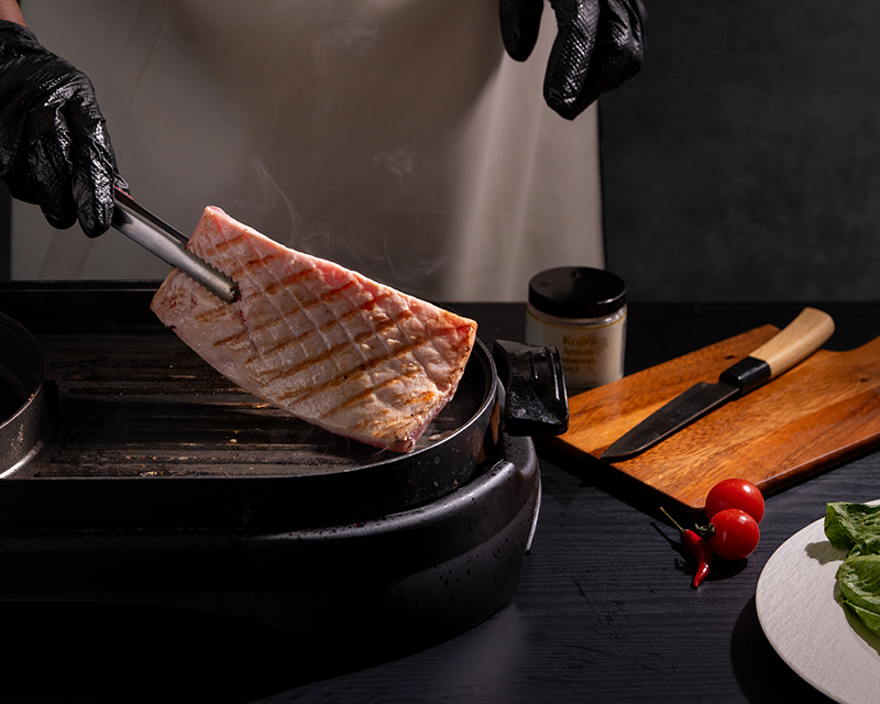 Tuna steak seasoning and searing on a hot grill pan, with a chef lifting the fish to reveal golden-brown grill marks.